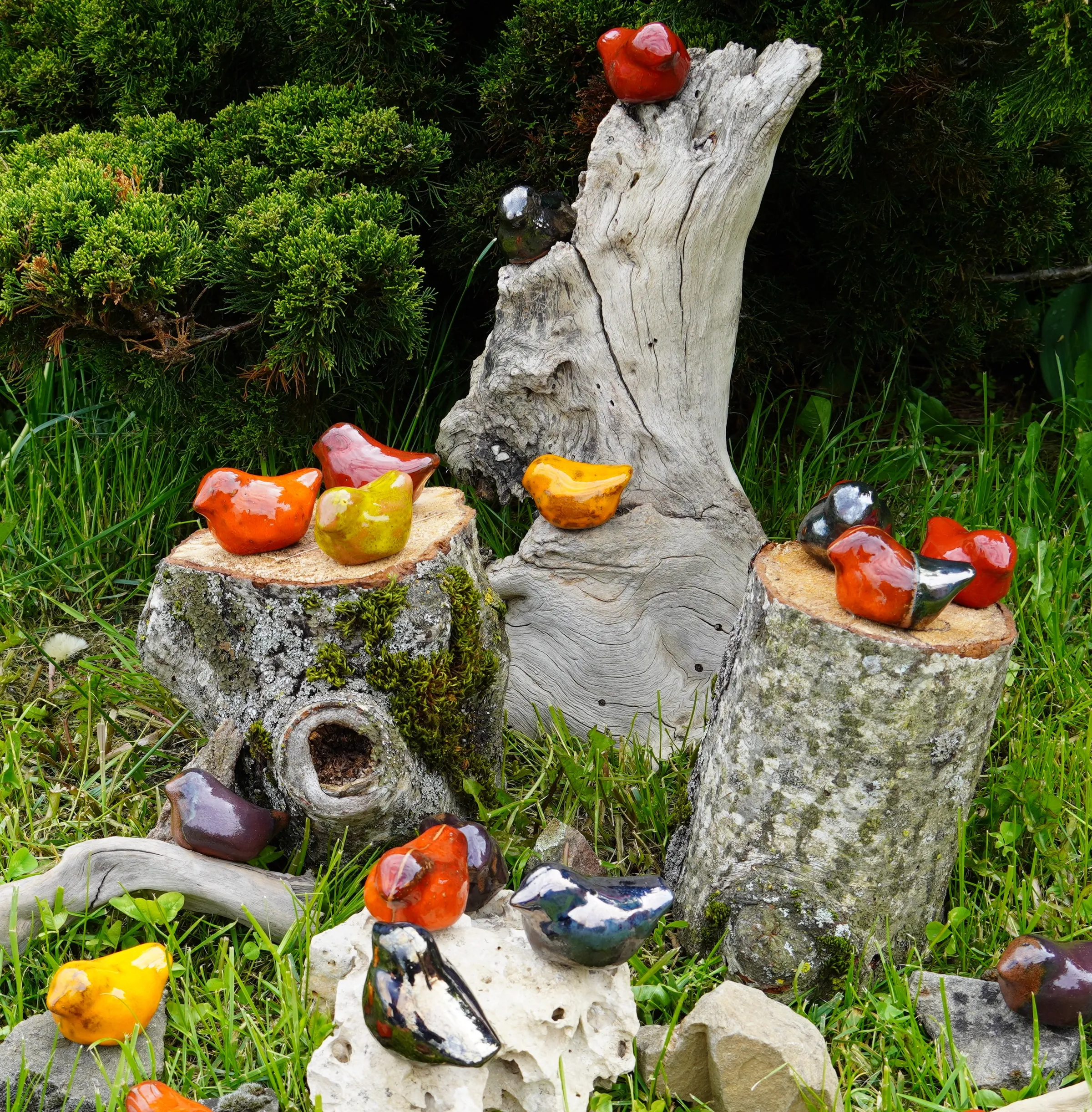 Ensemble d'oiseaux en céramique de differerentes couleurs, posés sur une buche dans l'herbe